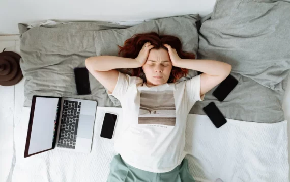 Woman surrounded by phones and laptop holding head